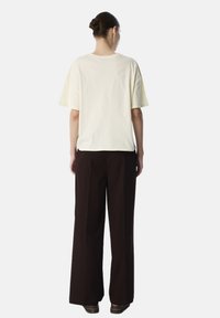 T-shirt giallo chiaro a maniche corte abbinato a pantaloni larghi marrone scuro, ripreso da dietro su uno sfondo minimal.