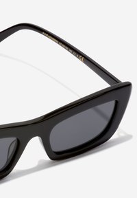 Svarta solglasögon med en djärv kattödesign, tillverkade av glänsande plast. Mörka linser och diskret guldbranding på den inre bygeln.