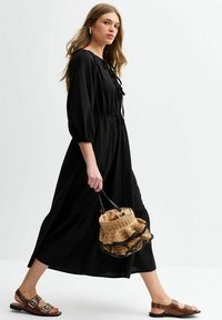 Black long-sleeve dress with a gathered waist and ruffle hem. Complemented by brown sandals and a woven bucket bag with a ruffled edge.