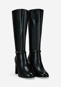 Un par de botas de cuero negro hasta la rodilla con tacones cuadrados, cremalleras laterales y correas en el tobillo con pequeñas hebillas de metal, exhibidas sobre un fondo blanco.