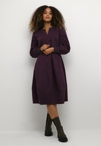 Ein langärmliges, knielanges Kleid in tiefem Lila mit Knopfdetails, das einen lockeren Schnitt und elastische Bündchen aufweist, getragen mit schwarzen klobigen Stiefeln.