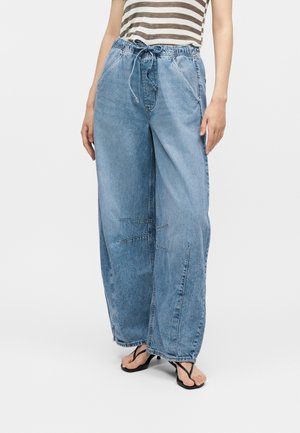 Άτομο που φοράει φαρδιά ανοιχτά γαλάζια τζιν παντελόνια με κορδόνι στη μέση, ριγέ αμάνικη μπλούζα και μαύρα σανδάλια με τακούνι, όρθιο.