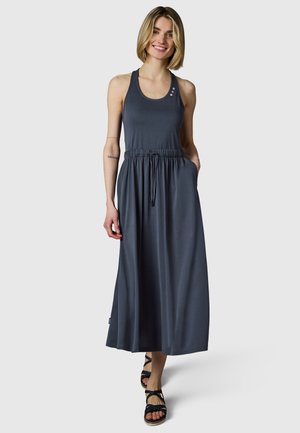 Hymyilevä nainen kävelee eteenpäin mustanharmaassa hihattomassa maksimekossa, jossa on nyöritys vyötäröllä, ja mustat remm sandaalit jalassa.