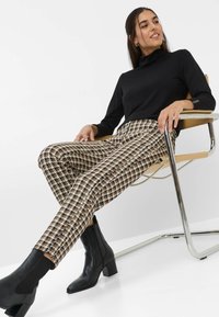 Zwarte lange mouwen top met een hoge kraag, gecombineerd met houndstooth-patronen tapered broek in bruin, crème en zwart; zwarte enkellaarsjes.