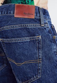 Primer plano de un bolsillo trasero de unos pantalones vaqueros azules con costuras amarillas y una etiqueta de cuero marrón de Pepe Jeans London.