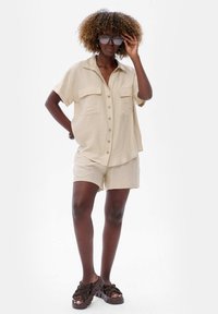 Beige linnen shirt met korte mouwen, voorzien van twee borstzakken en knoopsluiting, gecombineerd met bijpassende shorts en bruine chunky sandalen.