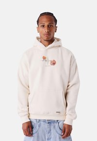 Sudadera crema con un bolsillo delantero, bordado floral en tonos durazno y rosa, capucha con cordones y puños de canalé. Llevada con jeans de color azul claro.