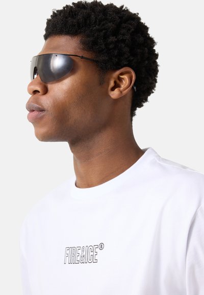 Jeune homme portant des lunettes de soleil enveloppantes réfléchissantes et un t-shirt blanc "FIRE & ICE", tourné vers la gauche sur un fond uni.