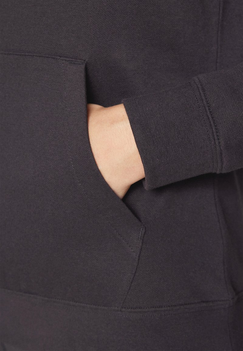 Dunkelgraues Sweatshirtmaterial mit einer Kängurutasche. Sichtbare Nähte und eine strukturierte Oberfläche. Hand, die in die Tasche gelegt ist.