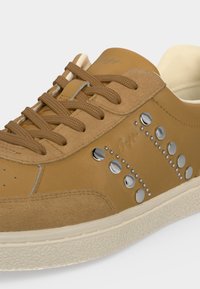 Baskets en cuir beige avec accents en daim, lacets marron, clous ronds argentés disposés en motif courbé, et semelle en caoutchouc blanche texturée.
