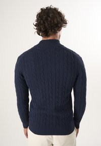 Dunkelblaues Strickpullover mit Kabelmustern, einem hohen Kragen sowie gerippten Bündchen und Saum. Die Textur wirkt weich und strukturiert.