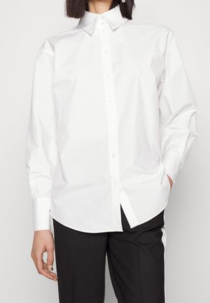 Femme portant une chemise blanche à manches longues boutonnée et un pantalon noir avec des bandes blanches sur les côtés, une main dans la poche.