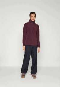 Νεαρός άνθρωπος που στέκεται φορώντας ένα μπορντό φλις με μισό φερμουάρ, χαλαρούς μαύρους παντελόνους και μωβ αθλητικά παπούτσια μπροστά σε έναν απλό ανοιχτό φόντο.