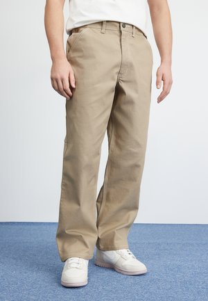 Man draagt beige wijde broekspijpen, witte sneakers en een wit overhemd, staand op een blauw tapijt tegen een effen lichte achtergrond.