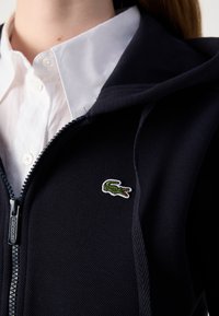 Sudadera con cremallera de color azul marino con tejido texturizado, que presenta un logo de Lacoste en blanco y un detalle bordado de cocodrilo verde en el pecho. Se vislumbra un cuello de camisa blanco.