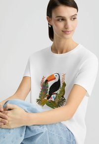 Balta trumpa rankovėmis marškinėliai su ryškiu tukano grafikų, apsuptu žalios augalijos ir gėlių raštų. Modelis sėdi, vilkėdamas šviesius džinsus.