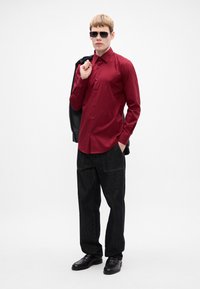 Uomo che indossa occhiali da sole e pantaloni neri, con una giacca nera sulle spalle, vestito con una camicia rossa a bottoni su uno sfondo bianco.