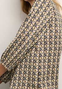 Mujer vestida con una blusa de patrón en beige, negro y blanco con diseños en forma de abanico y mangas de tres cuartos, mostrada de lado y por detrás.