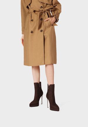 Trench color cammello fino al ginocchio con doppi bottoni e cintura in vita, abbinato a stivaletti marrone scuro con tacco alto e cerniere laterali.