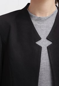 Musta takki, jossa on ainutlaatuinen viistonnappi suunnittelu, valmistettu sileästä materiaalista, käytetty vaaleanharmaan ribbipuseron päällä.
