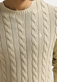 Pull en tricot beige avec un motif torsadé épais porté par une personne, montrant le cou et une partie du bras droit.