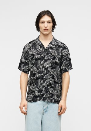 Młoda osoba o średniej długości ciemnych włosach, ubrana w czarną koszulę na krótki rękaw z białym wzorem liści oraz jasnoniebieskie dżinsy.