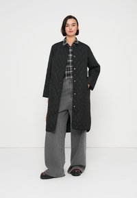 Μαύρο μακρύ καπιτονέ παλτό με κουμπιά τύπου σνάπ, με μοτίβο διαμαντιού και πλαϊνές τσέπες, φορεμένο πάνω από ένα πουκάμισο με καρό μοτίβο και φαρδιά γκρι παντελόνια.