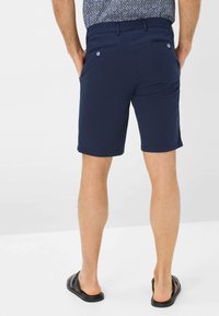 Dunkelblaue Shorts aus glattem Material. Mit zwei Gesäßtaschen mit Knopfverschluss, umgenähten Beinen und einem lockeren Schnitt, getragen mit schwarzen Riemensandalen.
