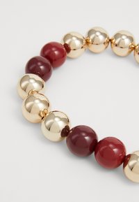 Armbånd med guld- og bordeauxfarvede perler, der har skinnende guldklodser og glatte bordeauxperler med indgraveret brandingdetalje.