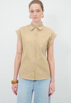 Beige Button-up-Hemd mit klassischem Kragen und hochgerollten kurzen Ärmeln; aus leichtem Stoff gefertigt; kombiniert mit hellblauen Jeans.