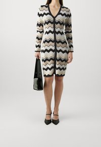 Robe en tricot avec un col en V, présentant un motif en zigzag en crème, marron et noir. Accompagnée d'un sac à main noir et de chaussures à talons pointues.