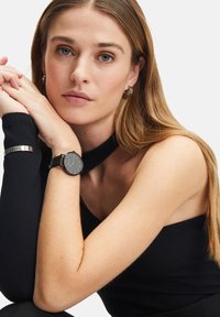 Schwarze Lederuhr mit einem einfachen runden Ziffernblatt, silbernen Markierungen und einem schwarzen gerippten ärmellosen Oberteil. Schlankes Design mit minimalem Zubehör.