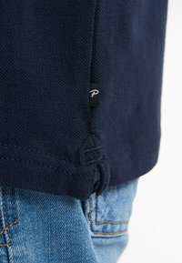 Mörkblå textured polo skjorta med en liten etikett vid fållen, tillsammans med ljusblå denimjeans. Visar detaljer kring sömmar och tyg.