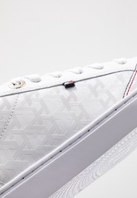 Białe sneakersy z teksturowanym wzorem geometrycznym, metalowymi oczkami i podwyższoną gumową podeszwą. Posiadają małe logo marki w kolorze czerwonym i czarnym.