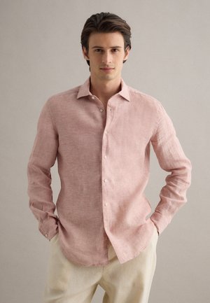 Hombre con camisa de manga larga color rosa claro y pantalones beige, de pie con las manos en los bolsillos frente a un fondo liso.