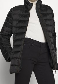 Svart vadderad puffjacka med dragkedjan öppen över en vit polotröja som är stoppad i högmidjade svarta byxor med synlig dragkedja och fickor.