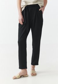 Pantaloni neri larghi con cintura a cravatta, gambe dritte e orli arrotolati, abbinati a sandali beige senza chiusura, su uno sfondo neutro.