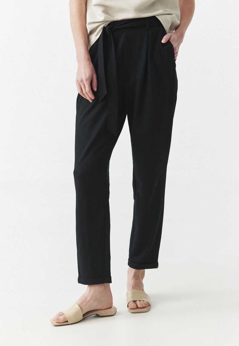 Pantaloni neri larghi con cintura a cravatta, gambe dritte e orli arrotolati, abbinati a sandali beige senza chiusura, su uno sfondo neutro.