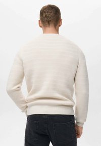 Suéter de punto color crema con un patrón texturizado, puños y dobladillo de canalé. Llevado con pantalones negros, visto desde atrás.