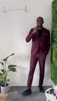 Traje burdeos con chaqueta de un solo boton y pantalones ajustados. Zapatos de vestir negros, micrófono en mano, planta en maceta al lado, pared verde como acento en el fondo.