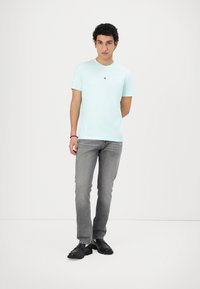 Hellblaues T-Shirt mit kurzen Ärmeln und kleinem Logo auf der Brust, kombiniert mit grauen Jeans und schwarzen Schnürschuhen.