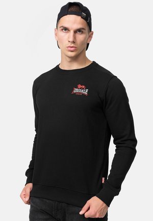 Sweat-shirt noir en tissu doux, avec un col rond. Logo rouge notable sur la poitrine et petite étiquette avec drapeau britannique sur le côté.