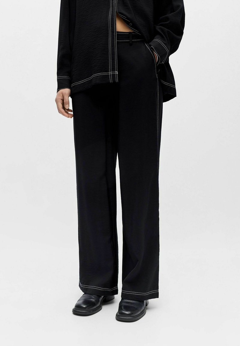 Personne portant un pantalon noir à jambes larges avec des coutures blanches, une chemise noire assortie et des chaussures noires sur un fond clair uni.