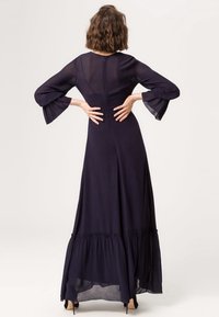 Robe longue bleu marine avec des manches cloche transparentes, un buste ajusté et un ourlet évasé avec une couture froncée. Détails de fermeture éclair au dos visibles.