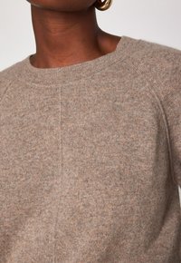 Maglione a maglia marrone chiaro con colletto rotondo, maniche raglan e una consistenza morbida, caratterizzato da sottili variazioni di colore in tutto il capo.
