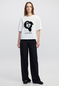 Witte T-shirt met korte mouwen heeft een zwart bloemmotief met een leeuwengrave. Gekoppeld aan zwarte wijde broek en versierde zwarte schoenen.