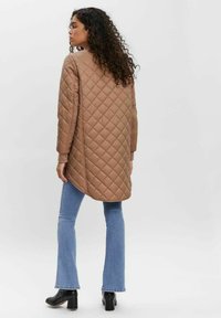Brown quilted long jacket with a rounded hem, ribbed cuffs, and a smooth texture, paired with light blue flared jeans and black heeled boots.