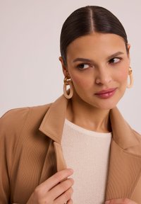 Beżowa, fakturowana kurtka z klapami, w zestawieniu z jasną, kremową, dzianinową bluzką. Modelka ma na sobie duże, okrągłe, jasne kolczyki. Zbliżenie.