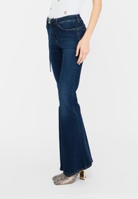 Femme portant un jean flare taille haute bleu foncé avec une ceinture fine et des bottines à talons en cuir serpent, se tenant devant un fond blanc.