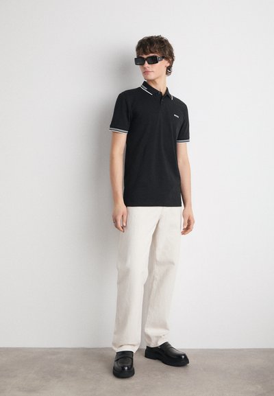 Polo noir avec des bordures blanches, associé à un pantalon beige clair et des chaussures noires. Modèle portant des lunettes de soleil sombres, se tenant devant un fond neutre.